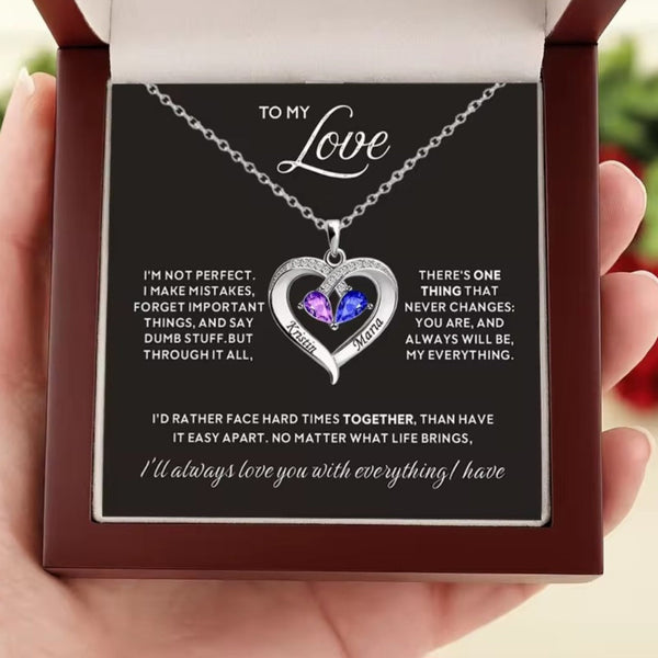 A heart-shaped silver necklace with a purple and blue gemstone center, presented inside a dark brown gift box held by a hand. The necklace is displayed on a black card with romantic and humorous quotes.